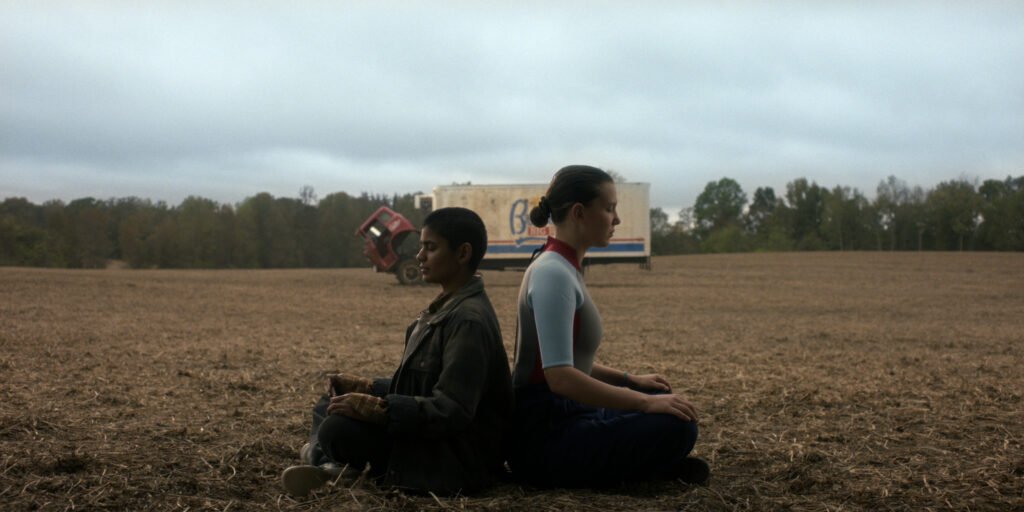 Kali e Eleven sentadas de costas uma para a outra em um campo aberto, em postura meditativa, sob um céu nublado e silencioso.