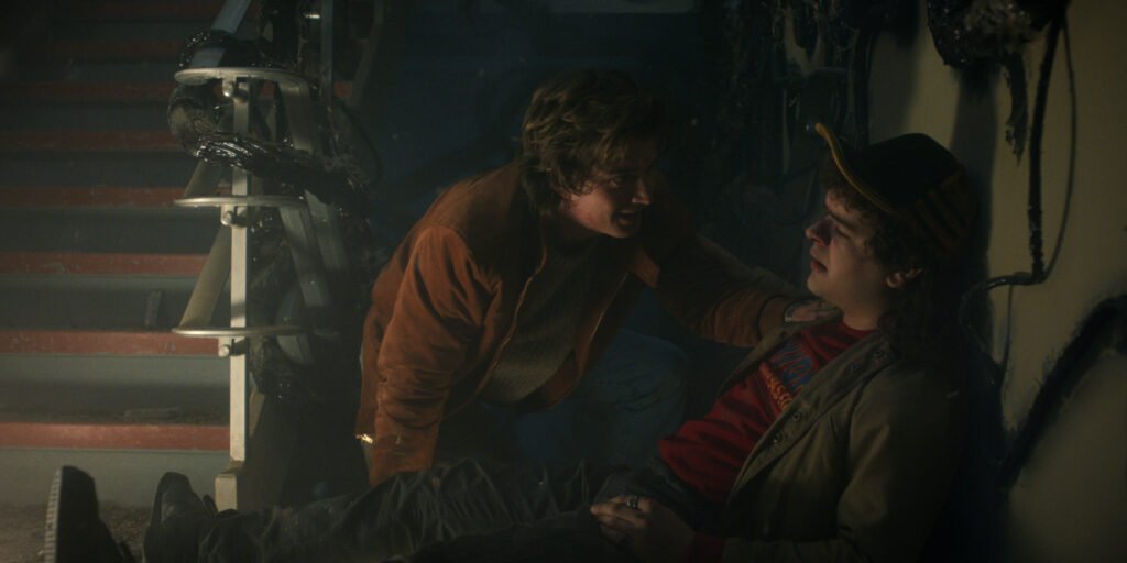Dustin e Steve em um ambiente escuro e danificado, onde um deles oferece apoio ao outro, em uma cena marcada por tensão e empatia.